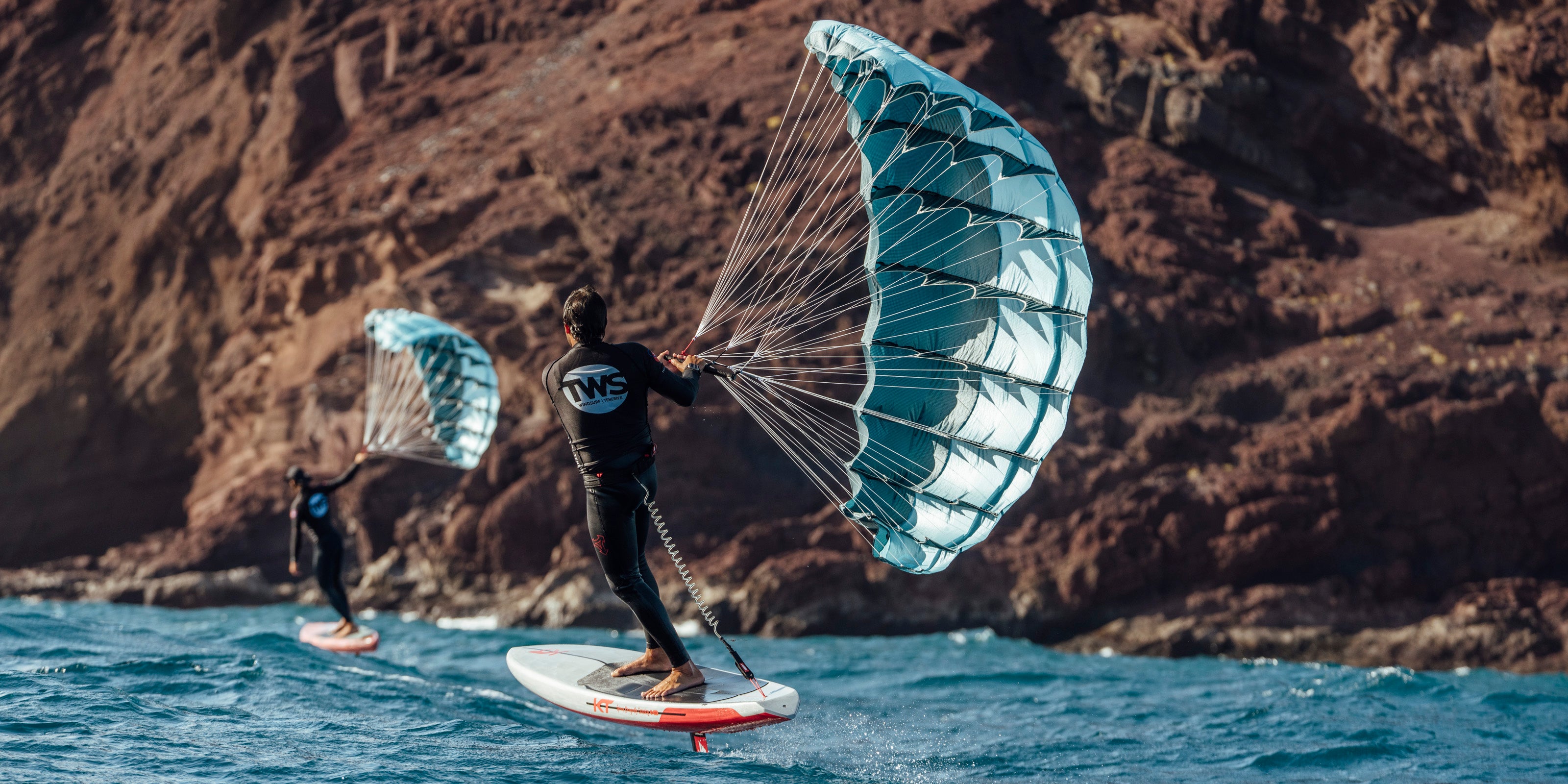 The setting appears to be coastal waters near rocky cliffs or mountains. The turquoise-blue wings stand out vividly against the brown/reddish backdrop of the terrain and the blue water. This relatively new watersport uses the wing to harness wind power while the hydrofoil beneath the board lifts the rider above the water's surface, reducing drag and allowing for smoother movement across the water.