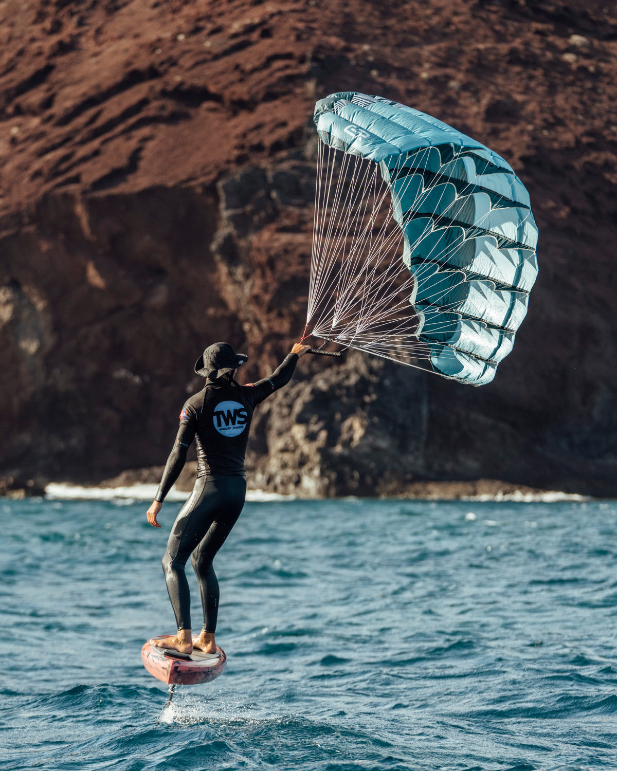 Turquoise Aeryn parawing lifts rider on foil board near rugged coastline. The wing's structured cells harness wind efficiently while white lines distribute force evenly. Hydrofoil technology elevates the board above water for minimal drag and smooth gliding. Perfect conditions showcase this innovative water sport's blend of surfing and wind power