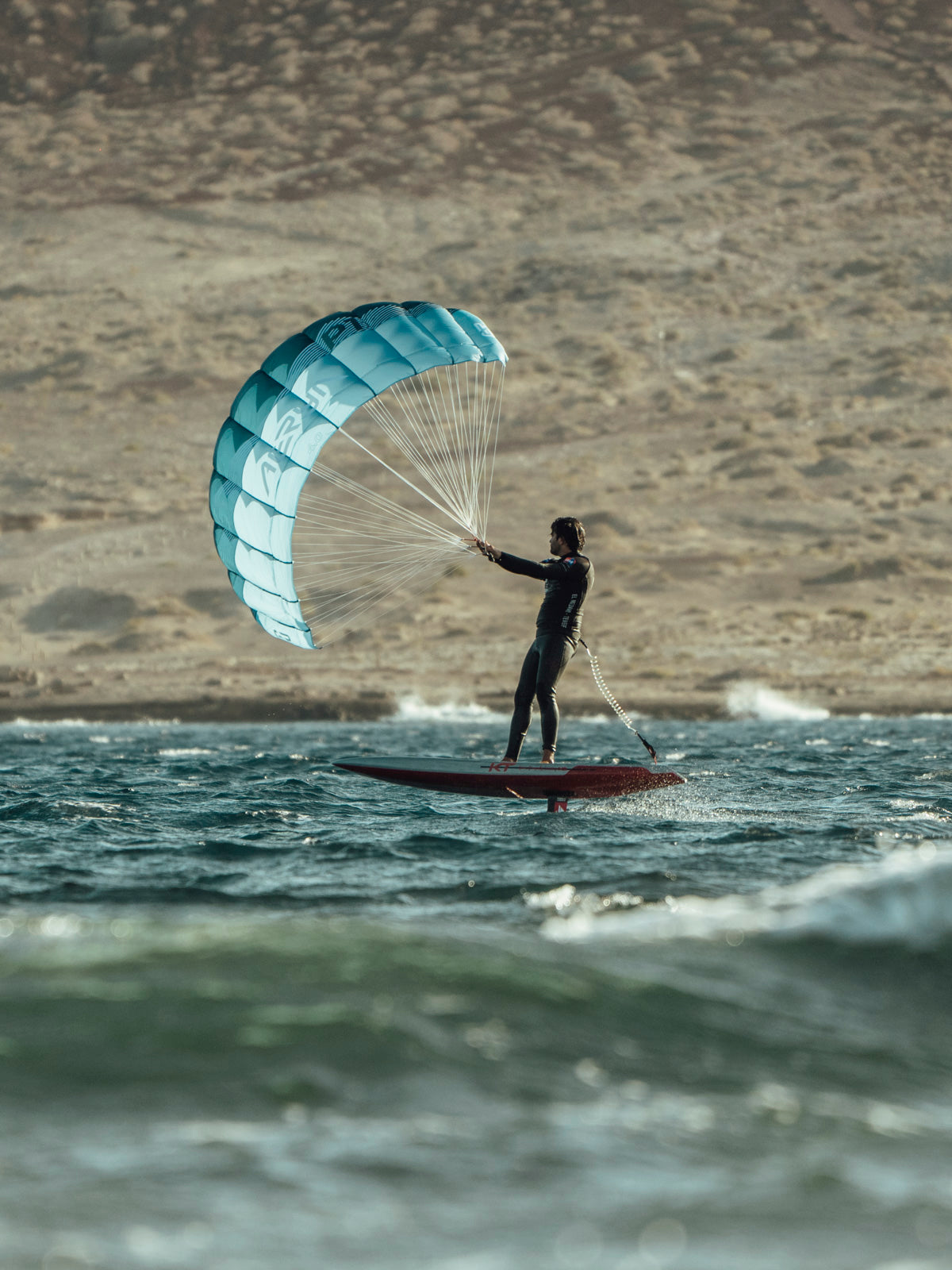 see a rider on a foil board with the distinctive turquoise aeryn parawing fully inflated against a desert/rocky coastal backdrop. The hydrofoil beneath the board is lifting the rider above the water's surface, demonstrating how the foil reduces drag for smoother movement.