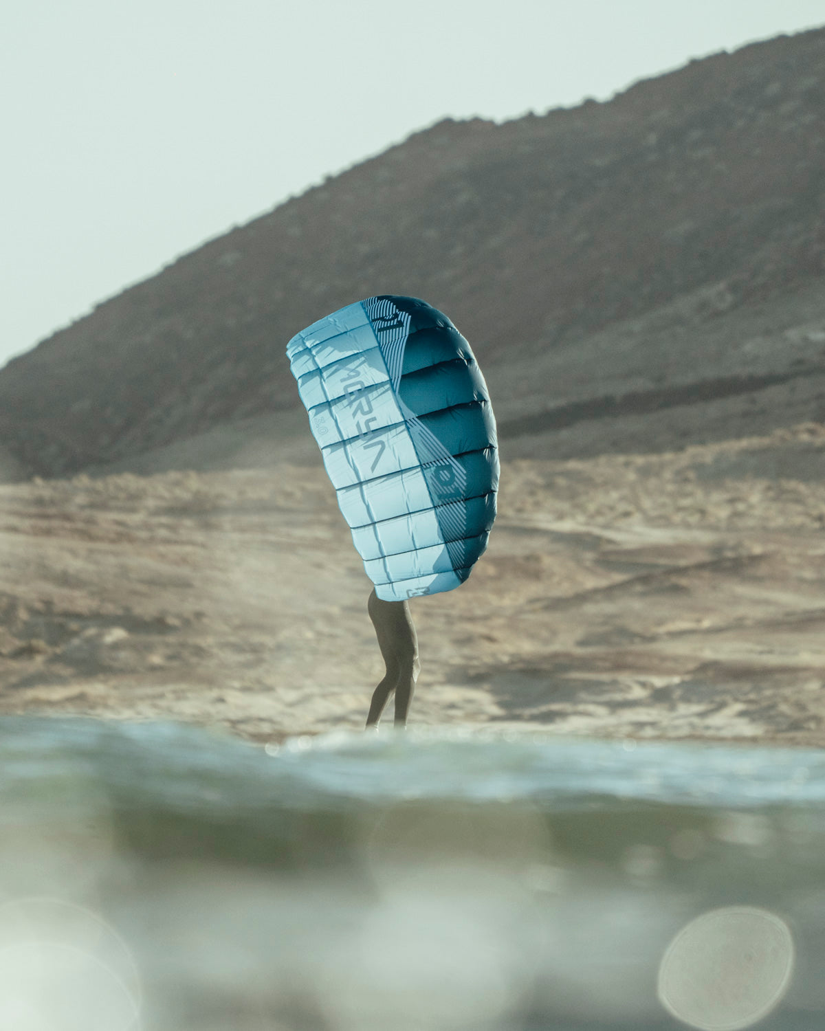Aeryn P1 pocket wing in action against desert backdrop. Turquoise parawing catches wind while foiler navigates coastal waters. The wing's structured cells and reinforced panels maintain perfect aerodynamic shape, showcasing its lightweight yet durable design for optimal performance.