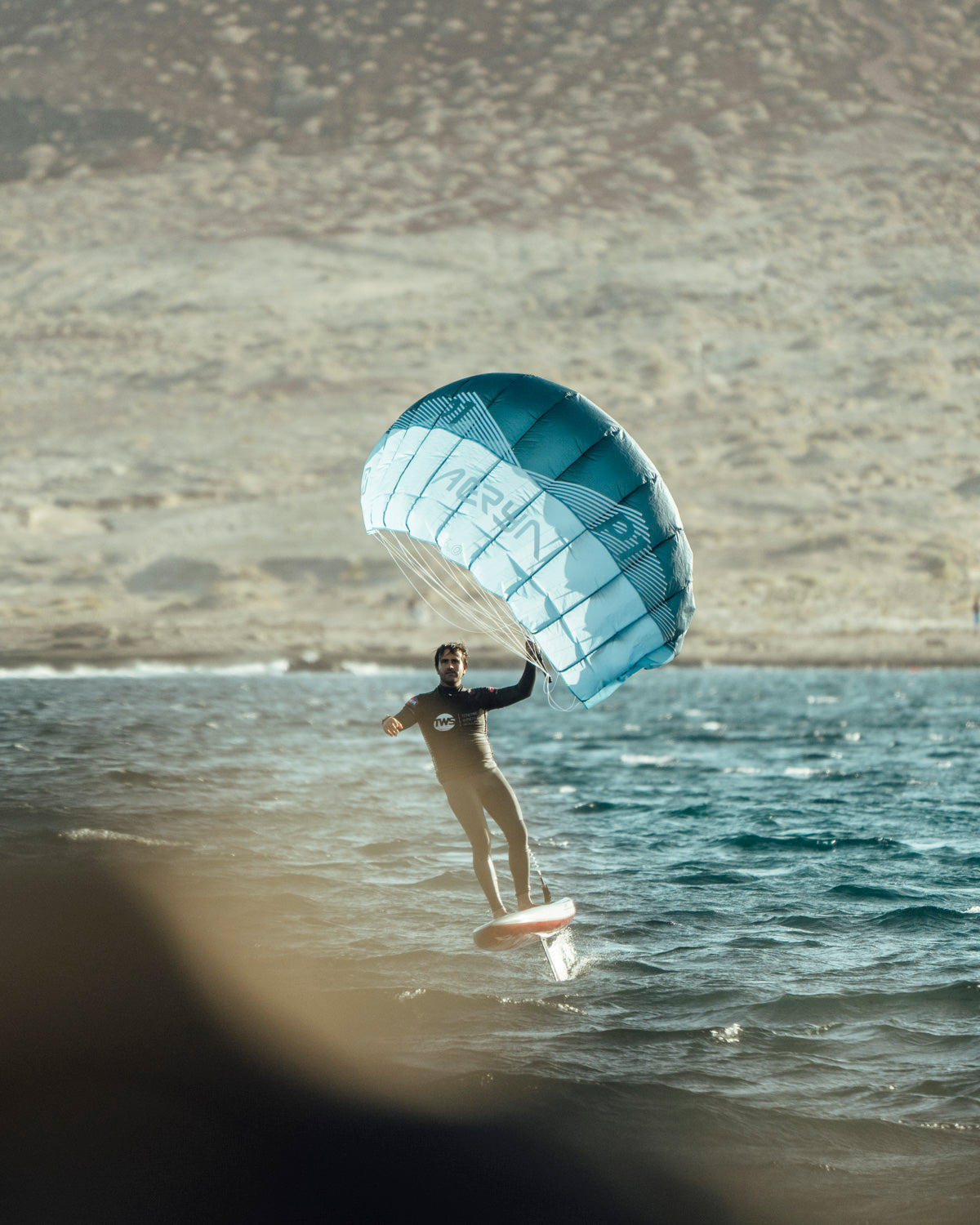 Aeryn P1 parawing elevates rider near shoreline. Turquoise wing captures wind as hydrofoil lifts board above water surface. Desert landscape contrasts with blue water, highlighting versatile locations for wing foiling. The wing's structured design with visible branding demonstrates professional-grade equipment in perfect flight position