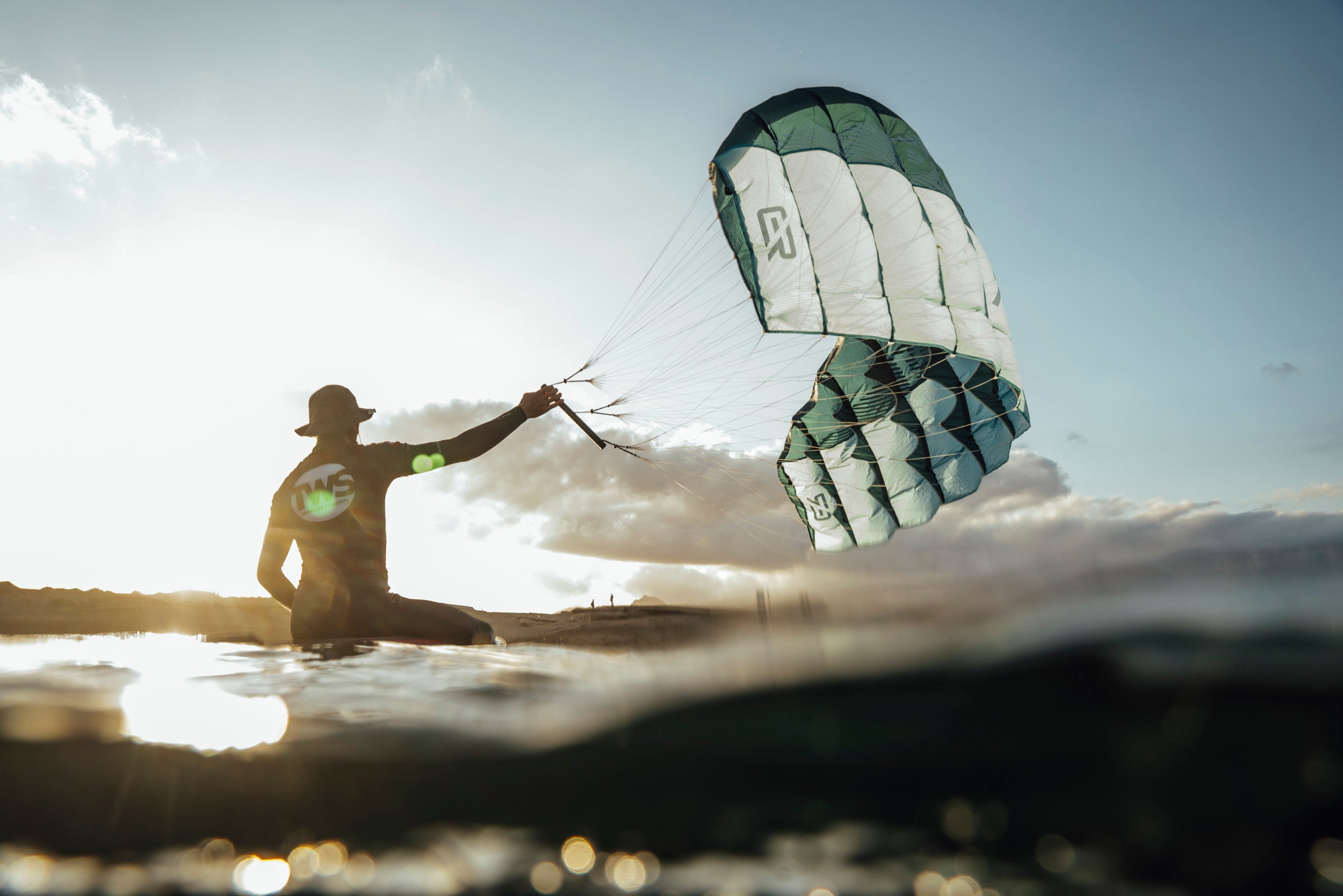 a rider sitting on a foil board and holding a small ram air kite that appear to be a parawing or pocket wing. The kite is blue and grey with a clear logo from Aeryn. the rider watch the sunset on his board