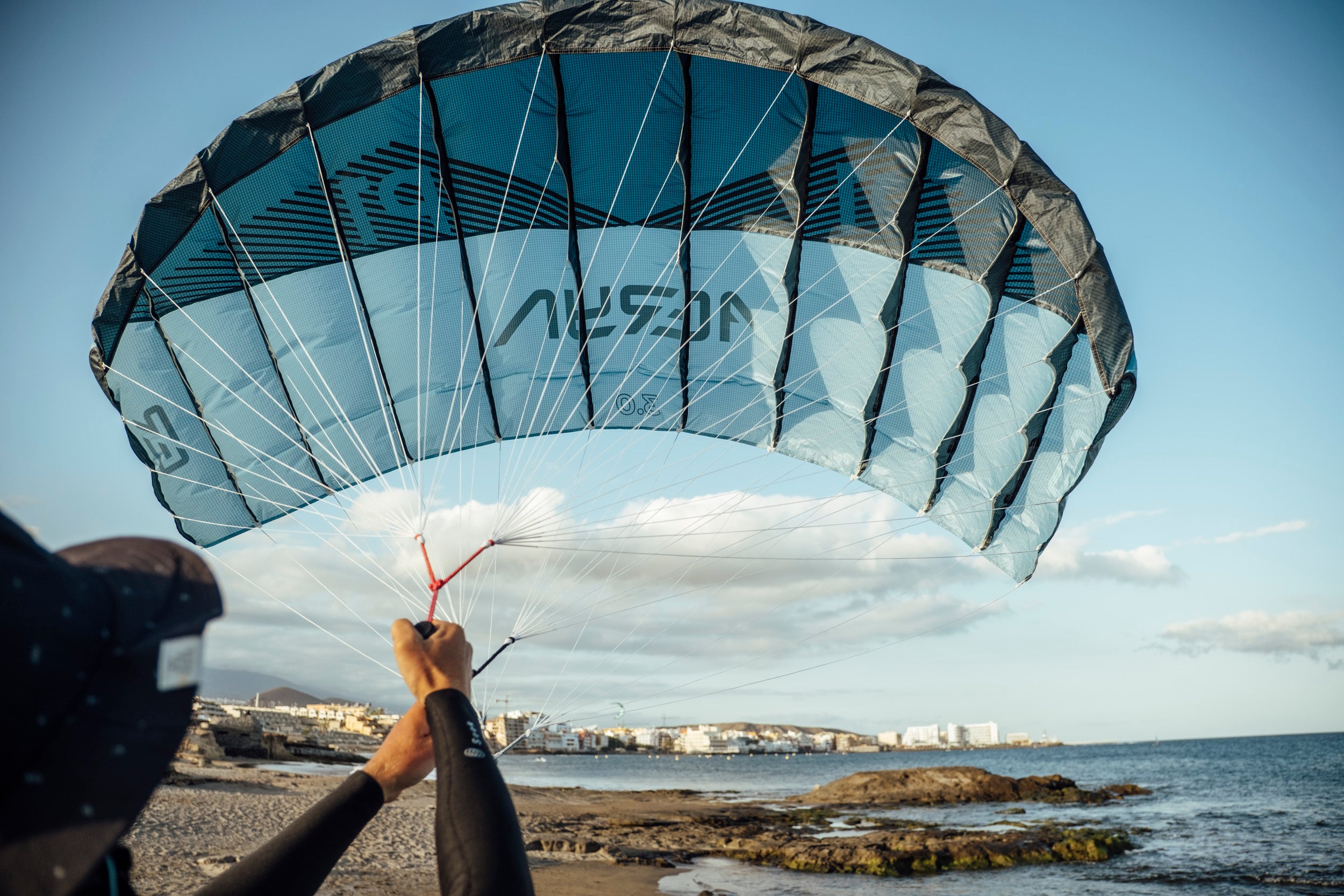 Aeryn parawing with distinctive blue and navy design fully deployed against clear sky. The wing displays "AERYN" branding across its reinforced panels. White control lines connect to the handlebar gripped from below. Coastal town visible in background showcases ideal foiling conditions with open water and steady winds. The structured cells and aerodynamic shape demonstrate premium Teijin fabric construction for optimal wind capture.
