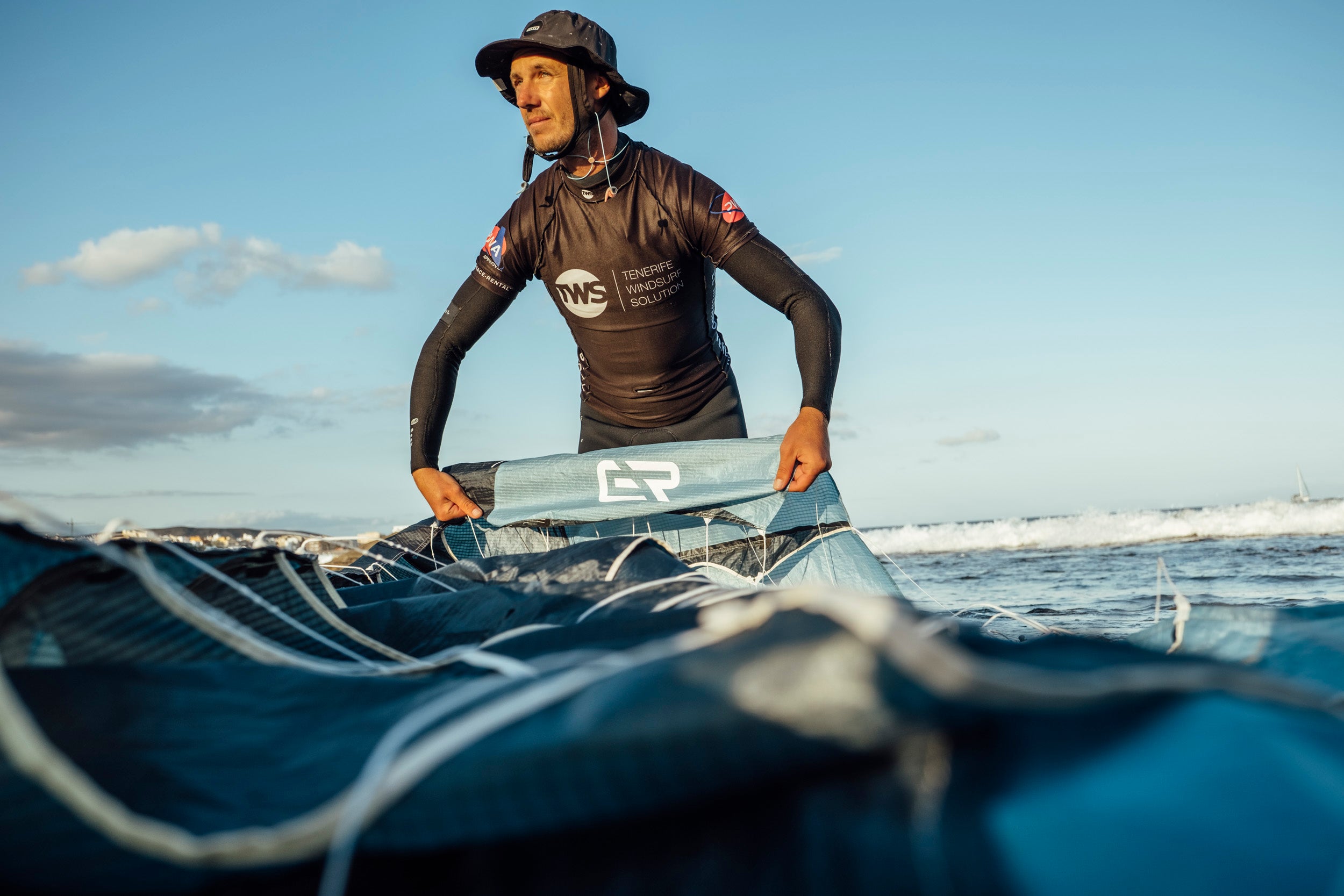 foiler preparing Aeryn parawing in water. Wearing protective wetsuit and hat, the rider handles the partially inflated blue wing with "TWS" logo visible. The shot from water level shows the wing's leading edge and structural lines as waves lap against it. Perfect conditions at Tenerife, known for consistent winds ideal for wing foiling, are evident with blue skies and gentle waves in the background.