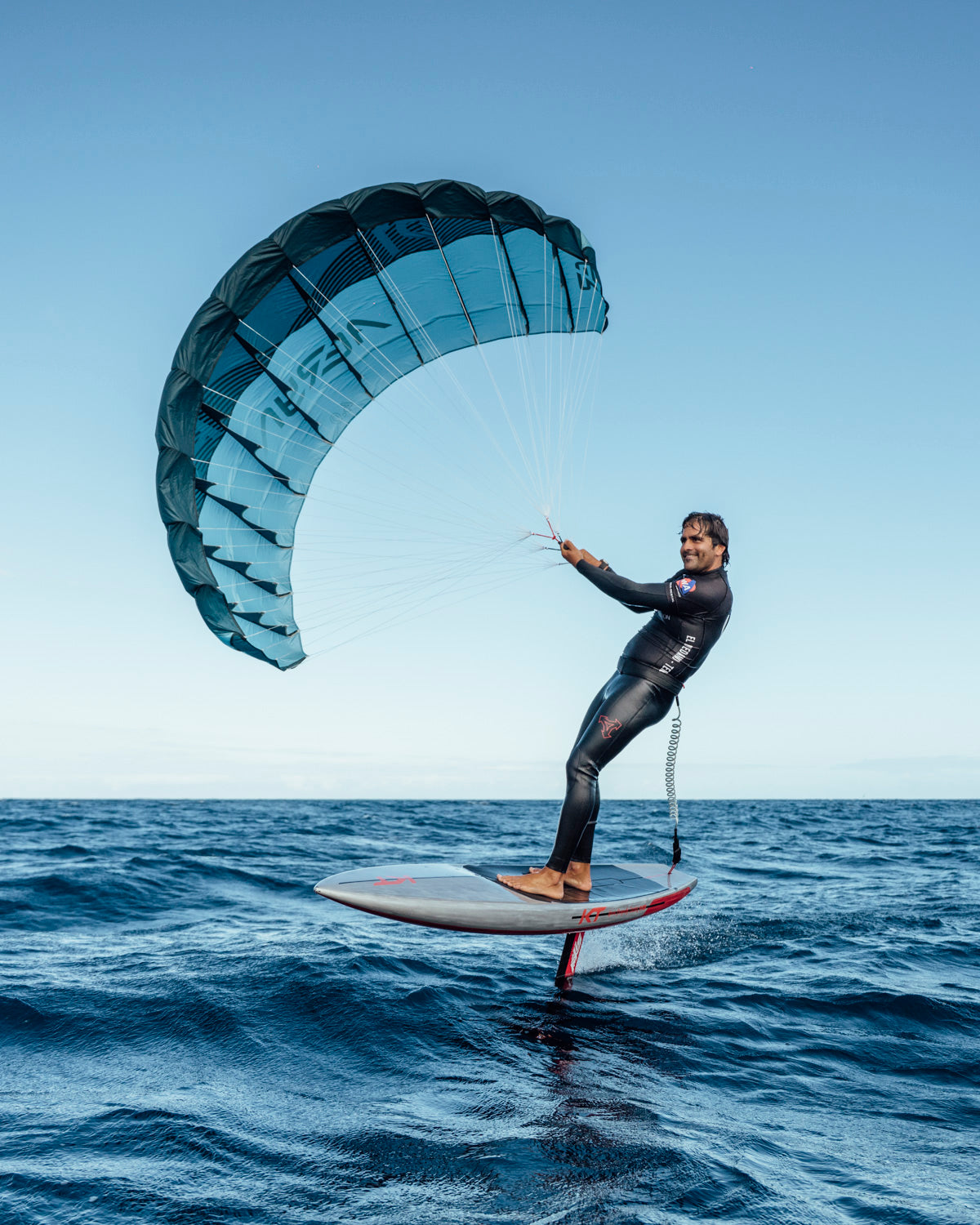 a rider in a black wetsuit seemingly floating above the water on his foil board. The turquoise and navy Aeryn P1 wing is fully deployed against a clear blue sky, showing the wing's perfect inflation and aerodynamic shape. The rider appears to be in mid-flight, showcasing the sport's dynamic quality.