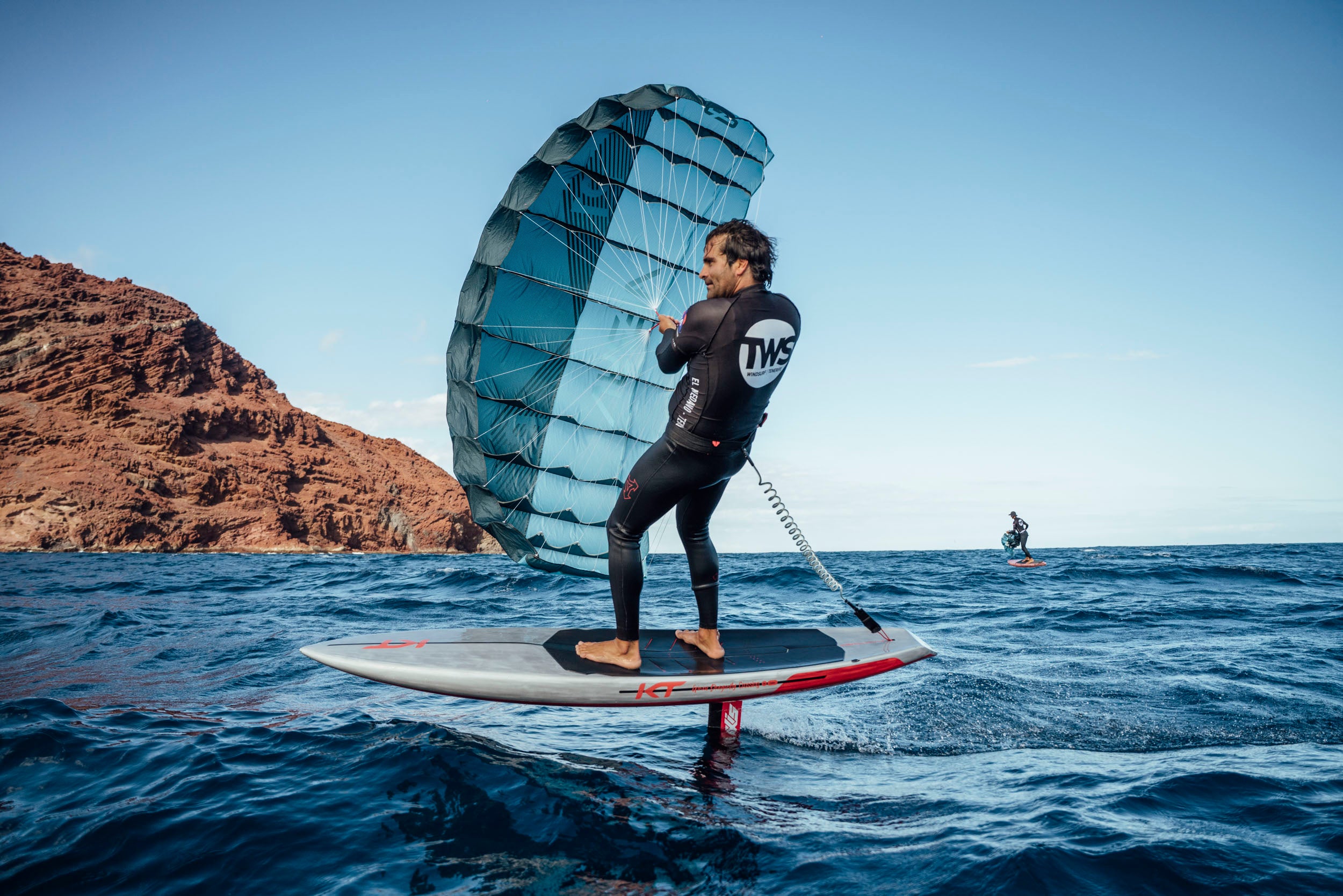 a rider on foil board holding a small kite with a small bar that appear to be a parawing or pocket wing. the wing is blue with a clear ocean background and a volcanic island that would appear to be tenerife