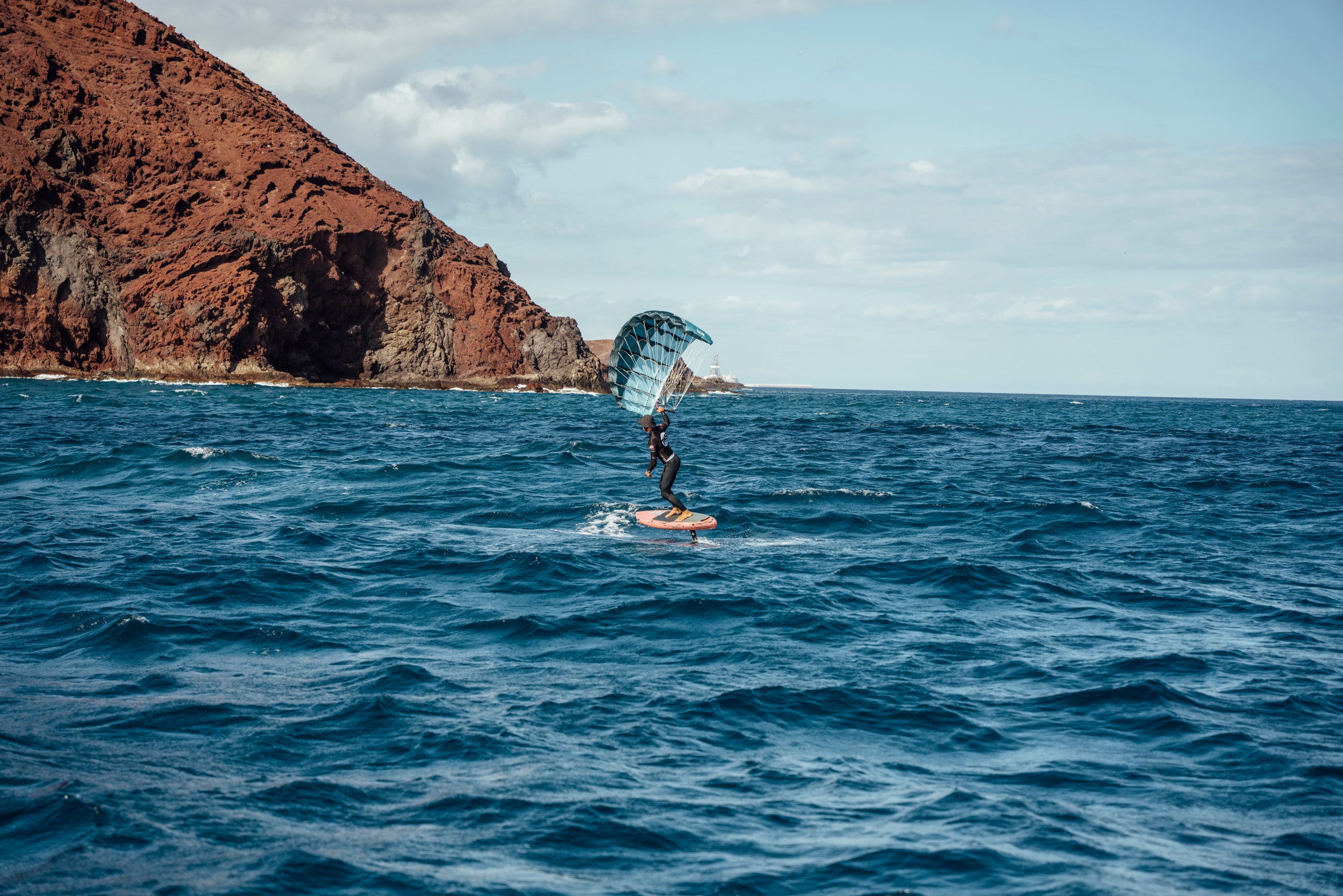 Aeryn pocket wing in action against dramatic red coastal cliffs. Turquoise parawing propels foil board across blue water, with hydrofoil lifting the rider above choppy waves. Perfect conditions showcase the sport's blend of wind power and water dynamics in this scenic natural setting.