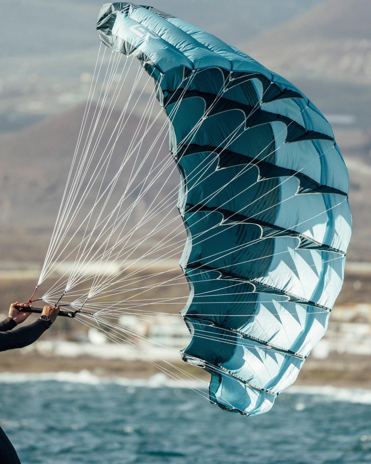 Aeryn P1 parawing showcasing its distinctive turquoise and navy pattern. The fully inflated wing displays the precision engineering with reinforced panels and carefully placed seams. White control lines connect to the handlebar, demonstrating the direct control system. The wing's aerodynamic shape captures wind efficiently for wing foiling, with coastal terrain visible in the background.