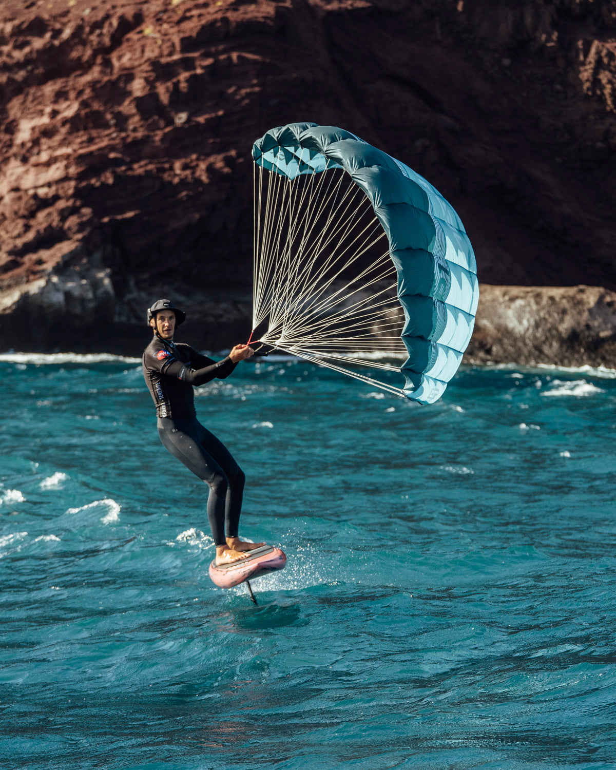 a wing foiler in coastal waters near dramatic rocky cliffs. The rider wears a cap while controlling the turquoise parawing. The white lines connecting the control bar to the wing are clearly visible, illustrating how the rider manages the wing's power and direction.