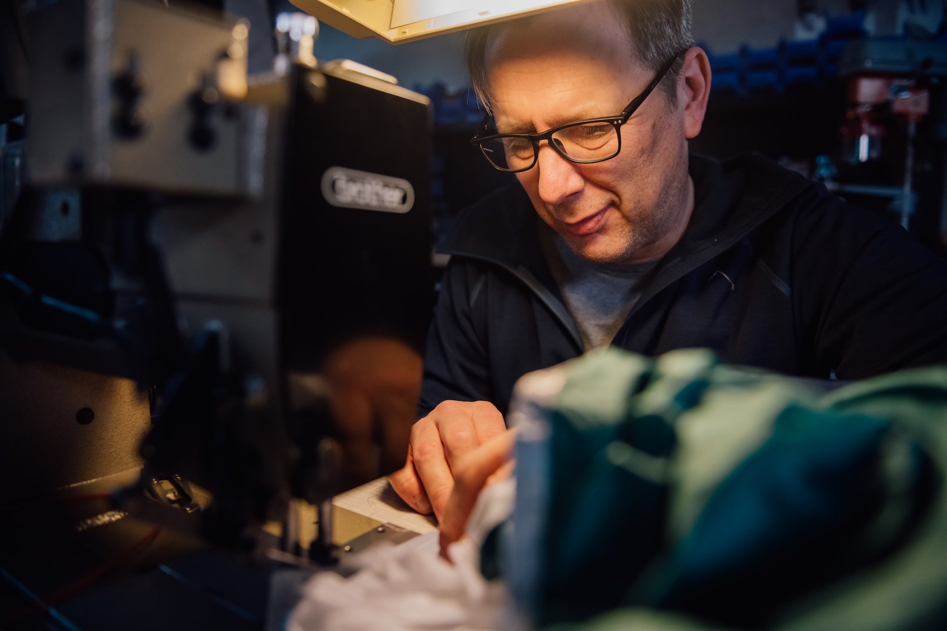 The image shows a craftsman at work, focused on the detailed process of creating wing foiling equipment. This appears to be Aeryn, a designer who works with PLKB (Peter Lynn Kiteboarding), carefully working on what might be a parawing or kite component. He's wearing glasses and a dark jacket, concentrating on a piece of material that's likely part of the parawing construction. The workshop environment shows specialized equipment for precision work, including what appears to be a sewing machine.