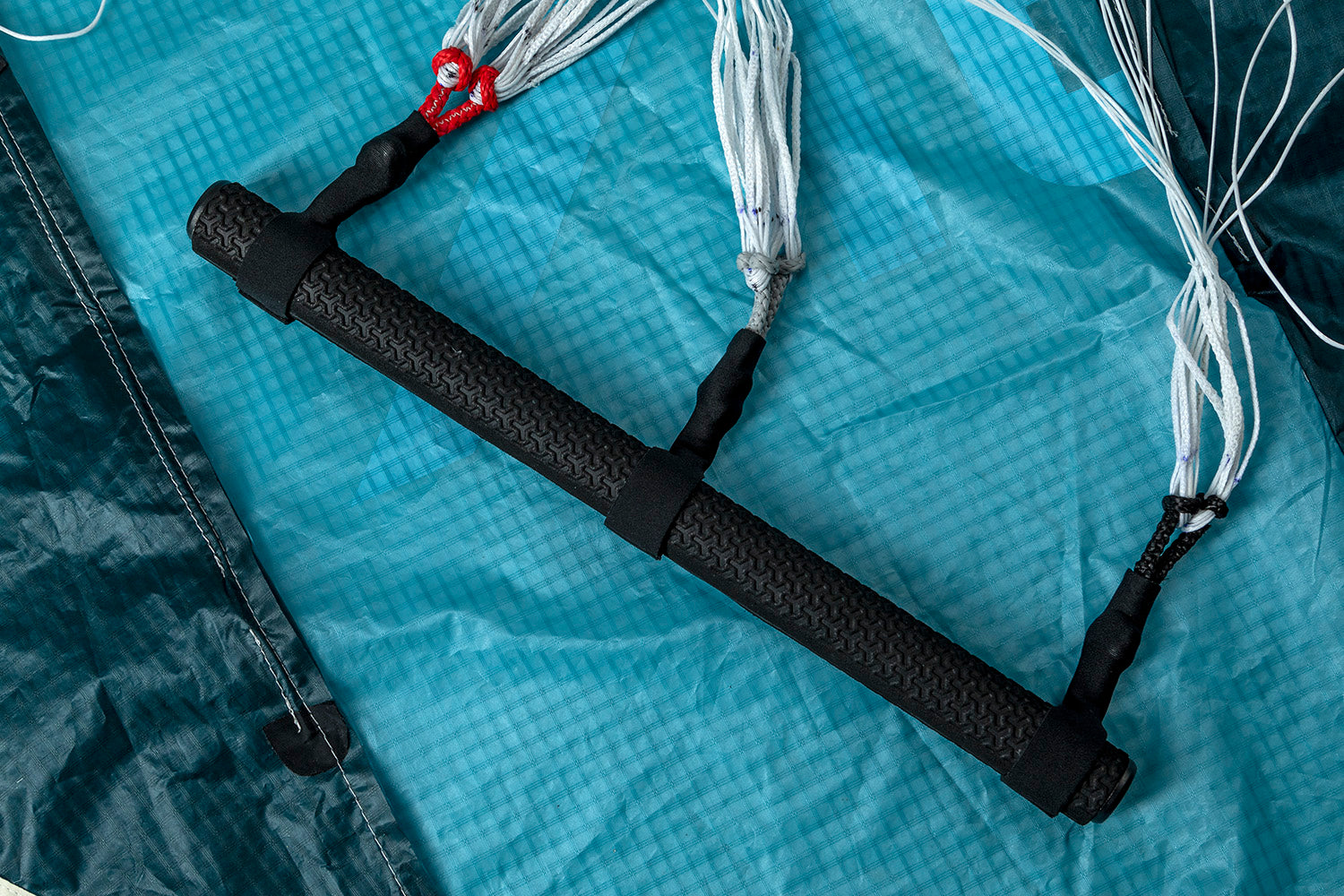 This is a control bar for wing foiling/wing surfing. The black textured handlebar connects to white lines/bridles with red loops visible on one end. It's placed on light blue ripstop fabric, likely part of the wing itself.