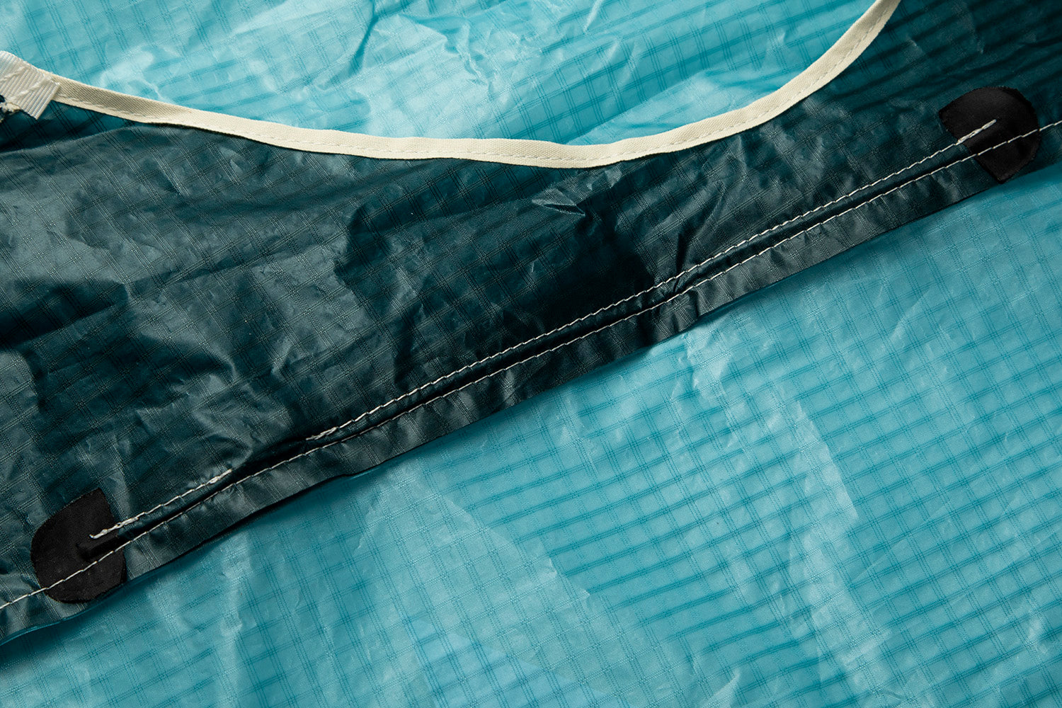 This image shows a detailed view of the parawing's material construction. The turquoise ripstop fabric makes up the main canopy, while a darker navy/black reinforced section with white stitching runs along a seam. Black rounded reinforcement patches are visible at stress points, likely where lines or other components attach. The beige/white trim at the top provides edge reinforcement. These quality construction details are essential for the parawing to maintain stability and durability when catching wind du