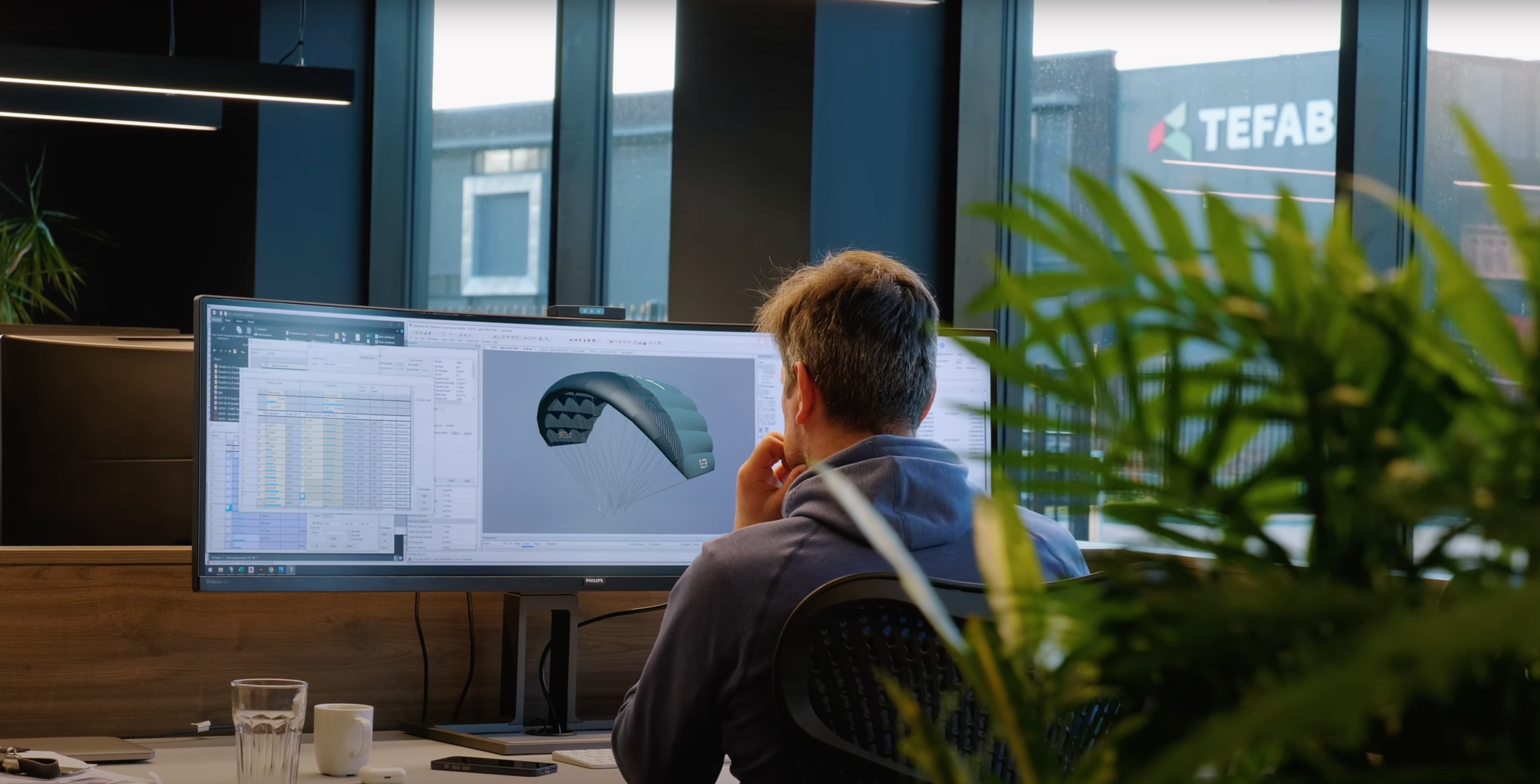 This image shows what appears to be Aeryn or another PLKB designer working on the digital modeling phase of parawing development. On the computer screen, a 3D rendering of a wing foiling parawing is visible alongside technical specifications or measurement data. The designer is seated at a modern office workstation, thoughtfully analyzing the design. This represents the high-tech aspect of modern wing development, where computer-aided design allows for precise modeling of aerodynamic properties