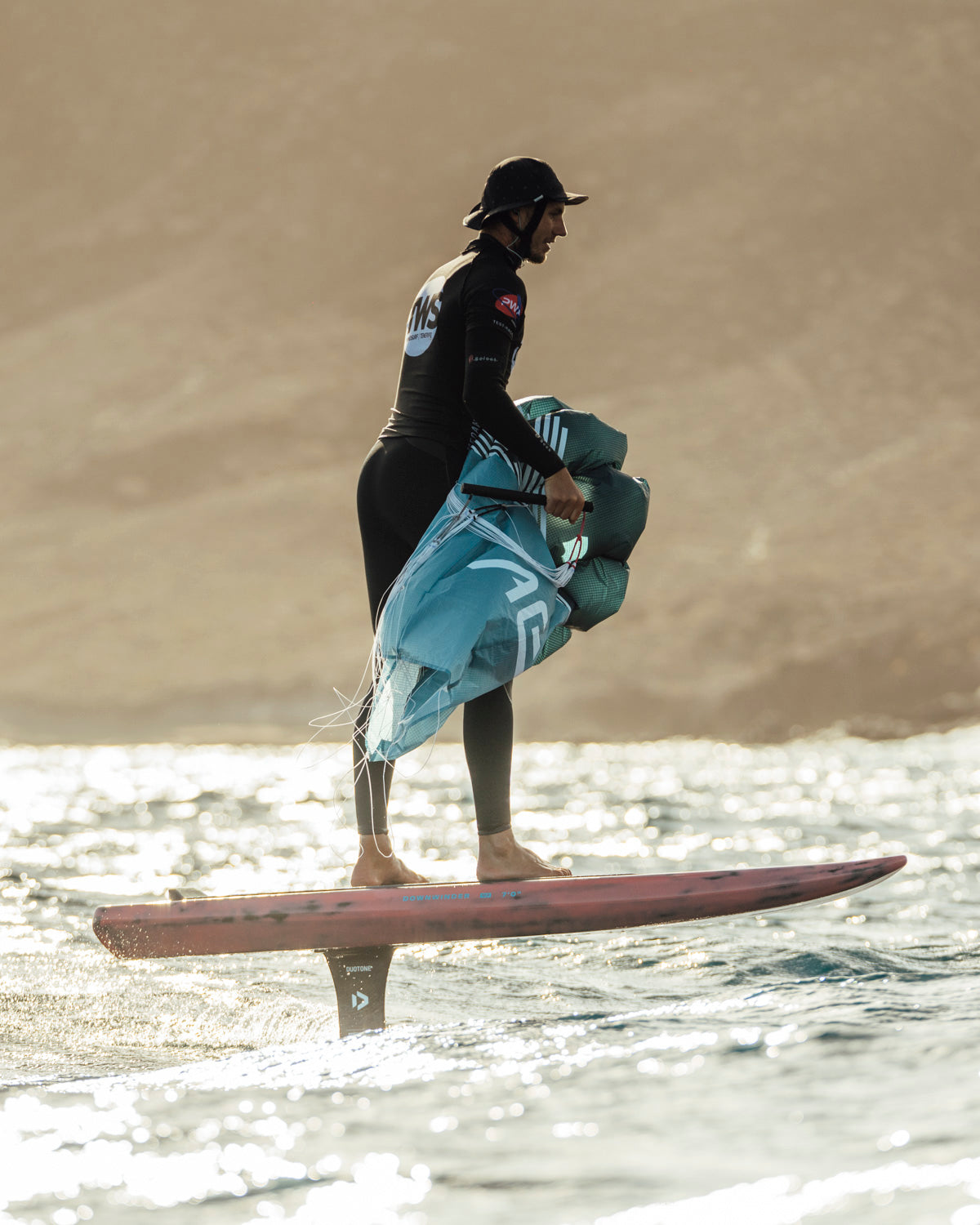 l  parawing ready for downwind action. The compact turquoise wing is being prepared on a red foil board at golden hour. The Duotone hydrofoil beneath provides lift for efficient gliding across water during downwind runs. Desert coastline backdrop highlights the versatile environments where this wing foiling equipment excels