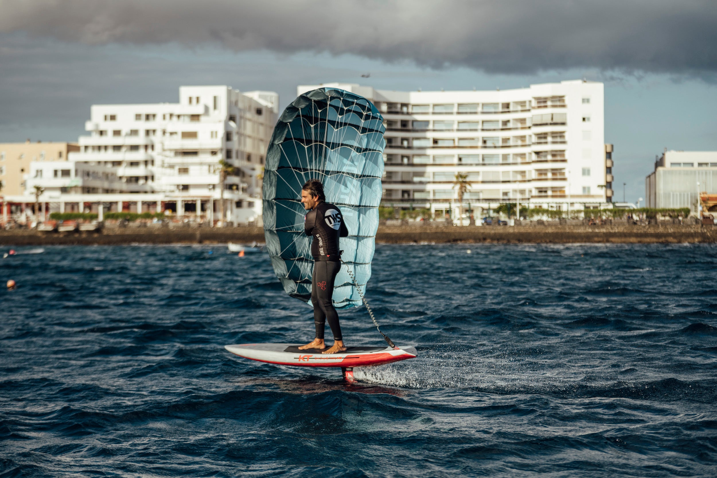 parawing in urban coastal action. A wing foiler stands on a hydrofoil board with the fully inflated blue wing catching wind. Beachfront buildings provide backdrop, demonstrating how this sport thrives in varied environments, from remote cliffs to developed waterfronts.